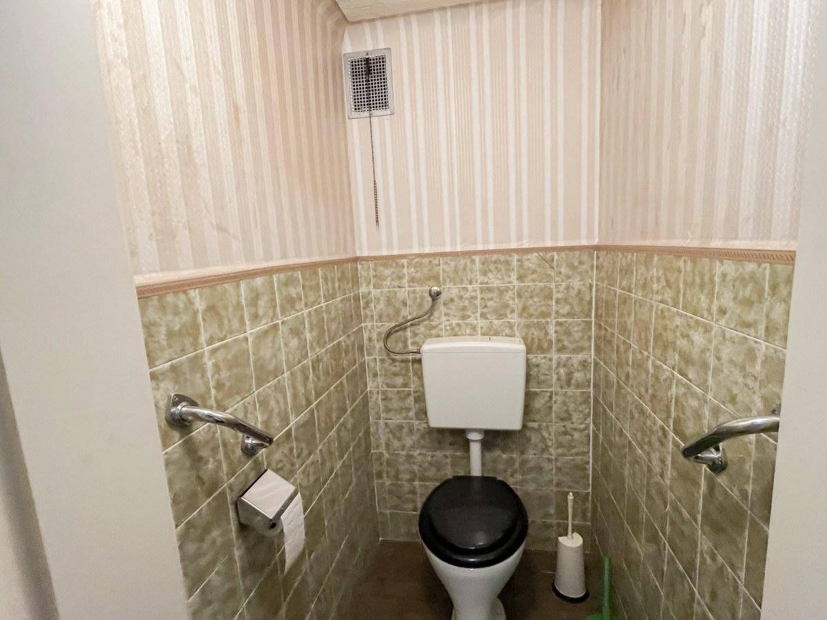 Kleines WC mit beigefarbenen Fliesen, gestreifter Tapete und Haltegriffen.