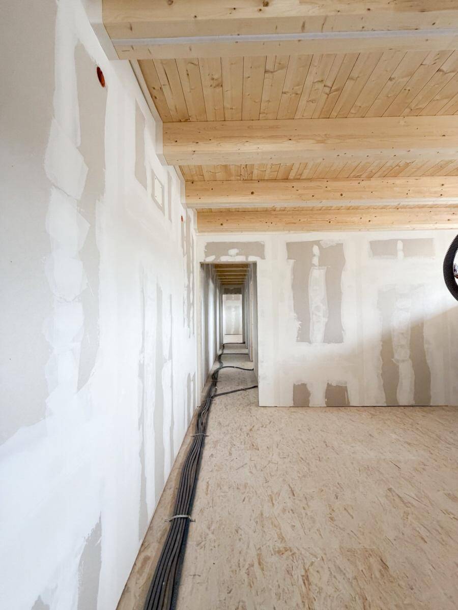 Langer Flur im Rohbau mit sichtbaren Holzbalken an der Decke und vorbereiteten Wänden für den Ausbau.