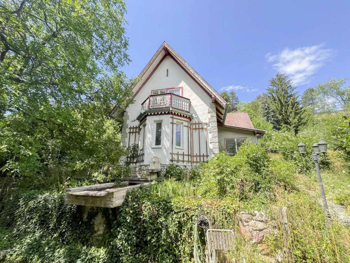 Charmantes altes Haus mit Balkon, umgeben von üppiger Vegetation und einem Brunnen.