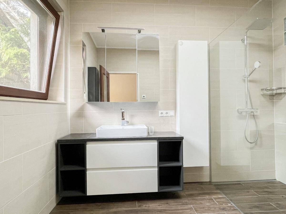 Badezimmer mit Fenster, modernem Waschtisch, begehbarer Dusche und hellen Wandfliesen.