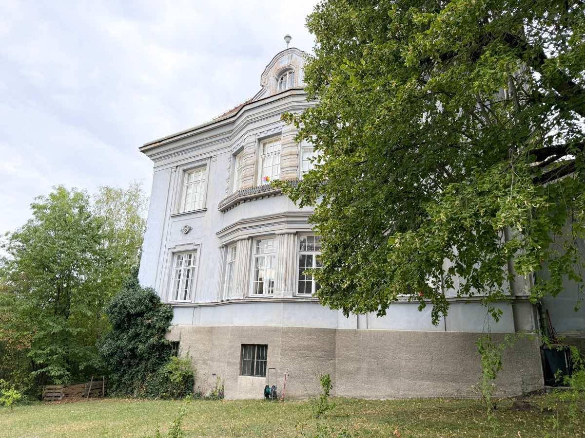 Historisches Gebäude mit klassischer Architektur und großen Fenstern, umgeben von Grün.