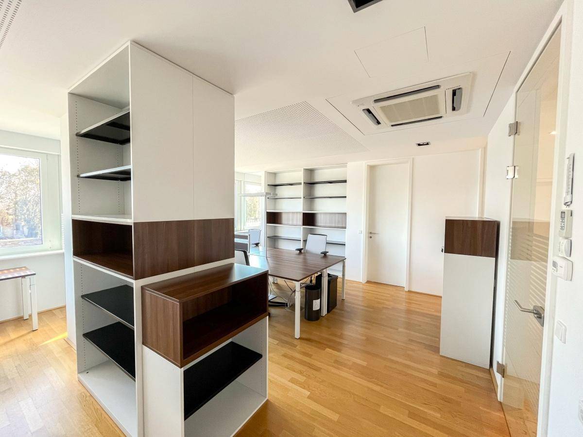 Geräumiges Büro mit Holzfußboden, weißen Regalen und integrierter Klimaanlage, bietet eine angenehme Arbeitsatmosphäre.