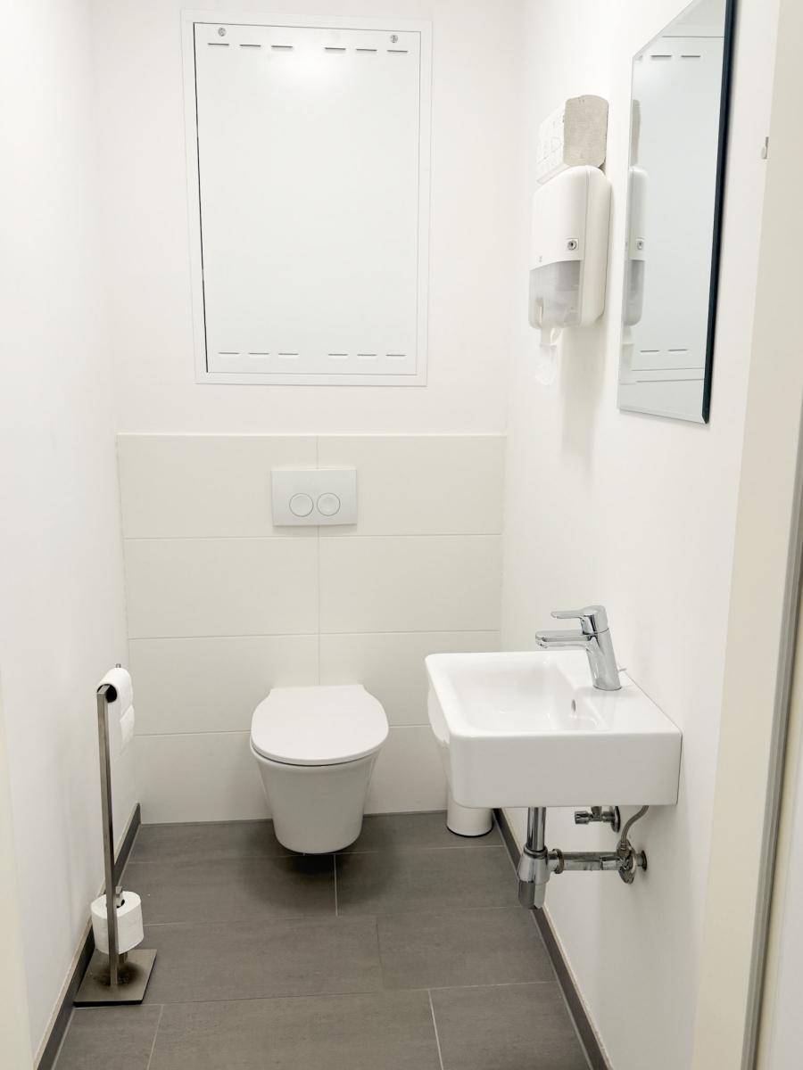 Helles Gäste-WC mit wandhängender Toilette, kleinem Waschbecken und grauen Fliesen.