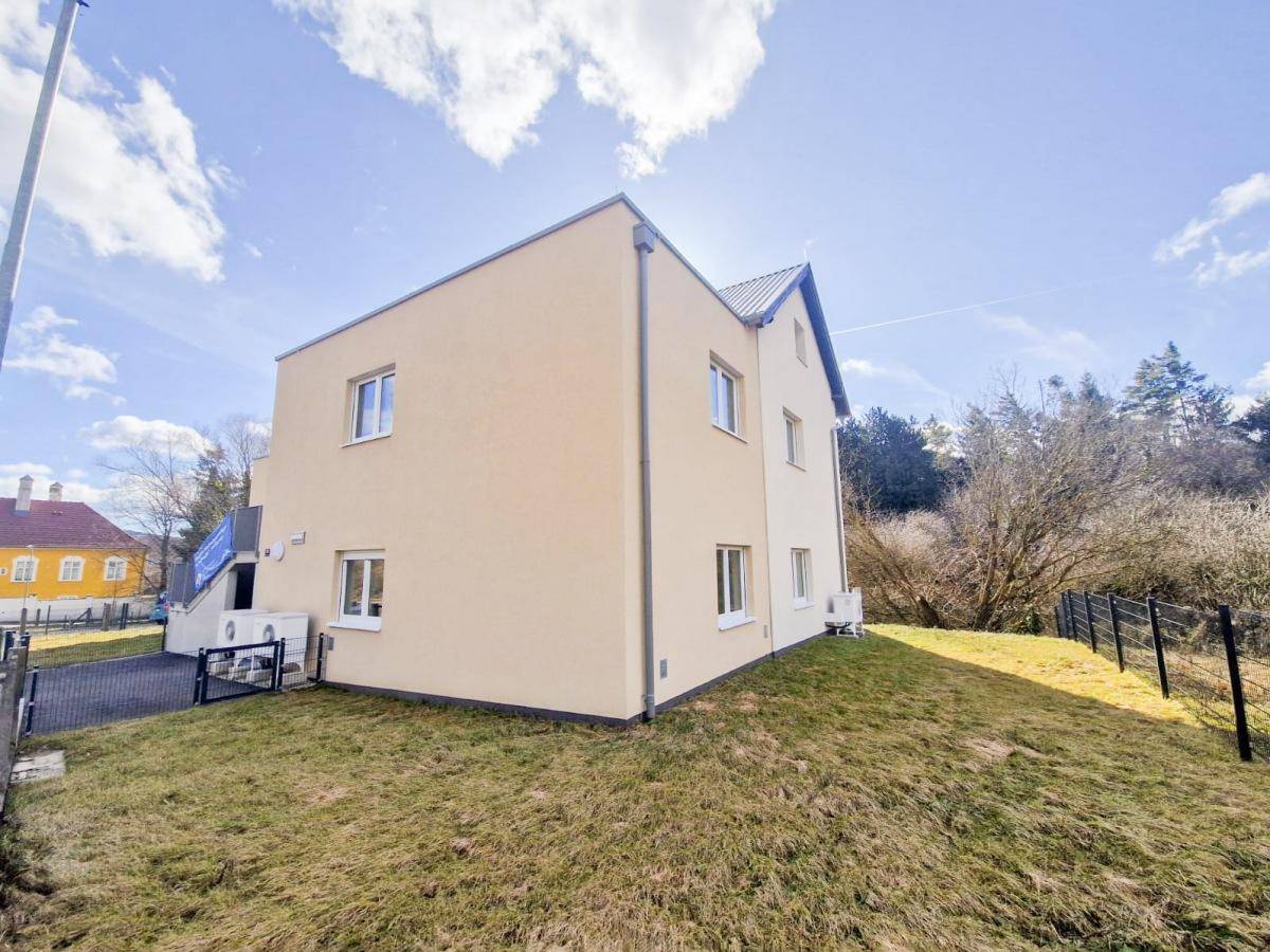 Seitenansicht des Neubaus mit beiger Fassade, Fenstern und einer gepflegten Rasenfläche.