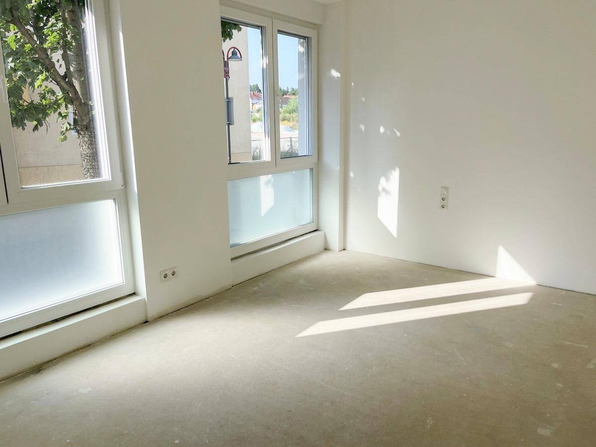Sonnenlicht durchflutet die Ecke eines leeren Zimmers mit großen Fenstern und Blick ins Grüne.