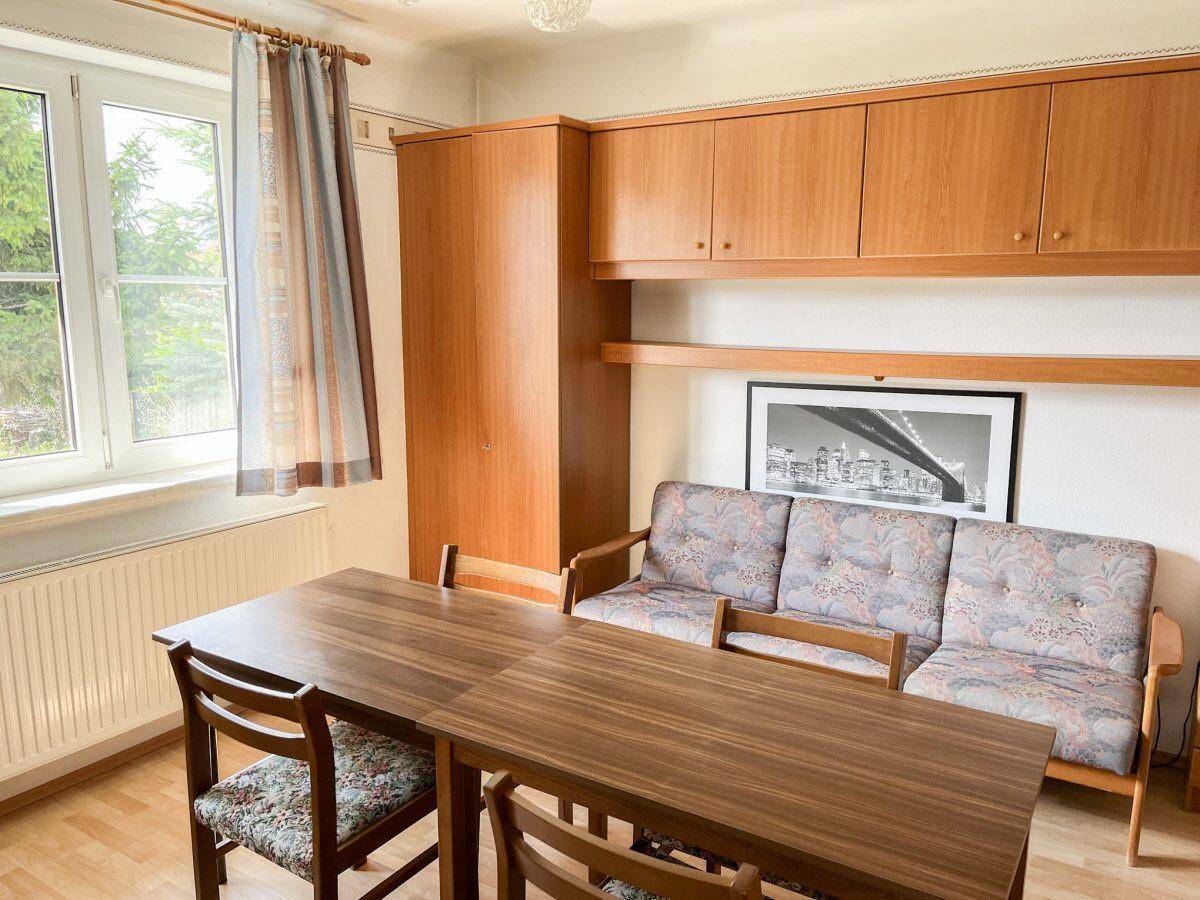 Gemütliches Wohnzimmer mit Holztisch, Sofa und Schrankwand, hell durch großes Fenster.