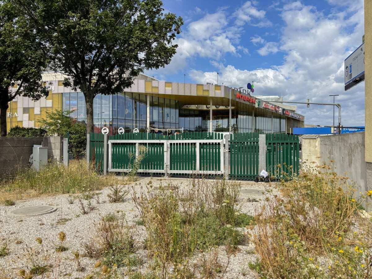 Blick vom Grundstück auf den grünen Zaun und ein großes Einkaufszentrum im Hintergrund.