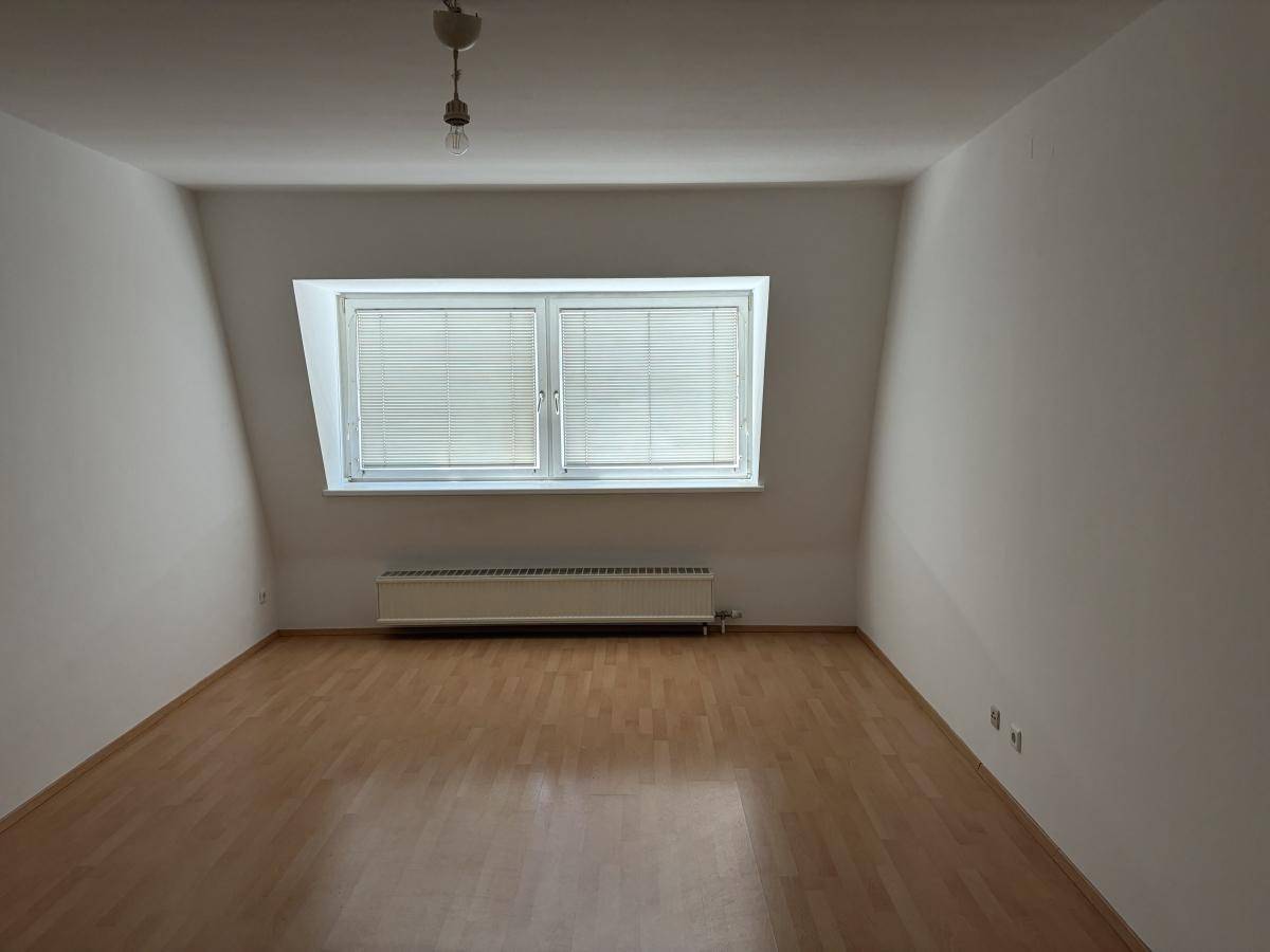 Leerer Raum mit Dachschrägen, großem Fenster und Laminatboden, gut beleuchtet.