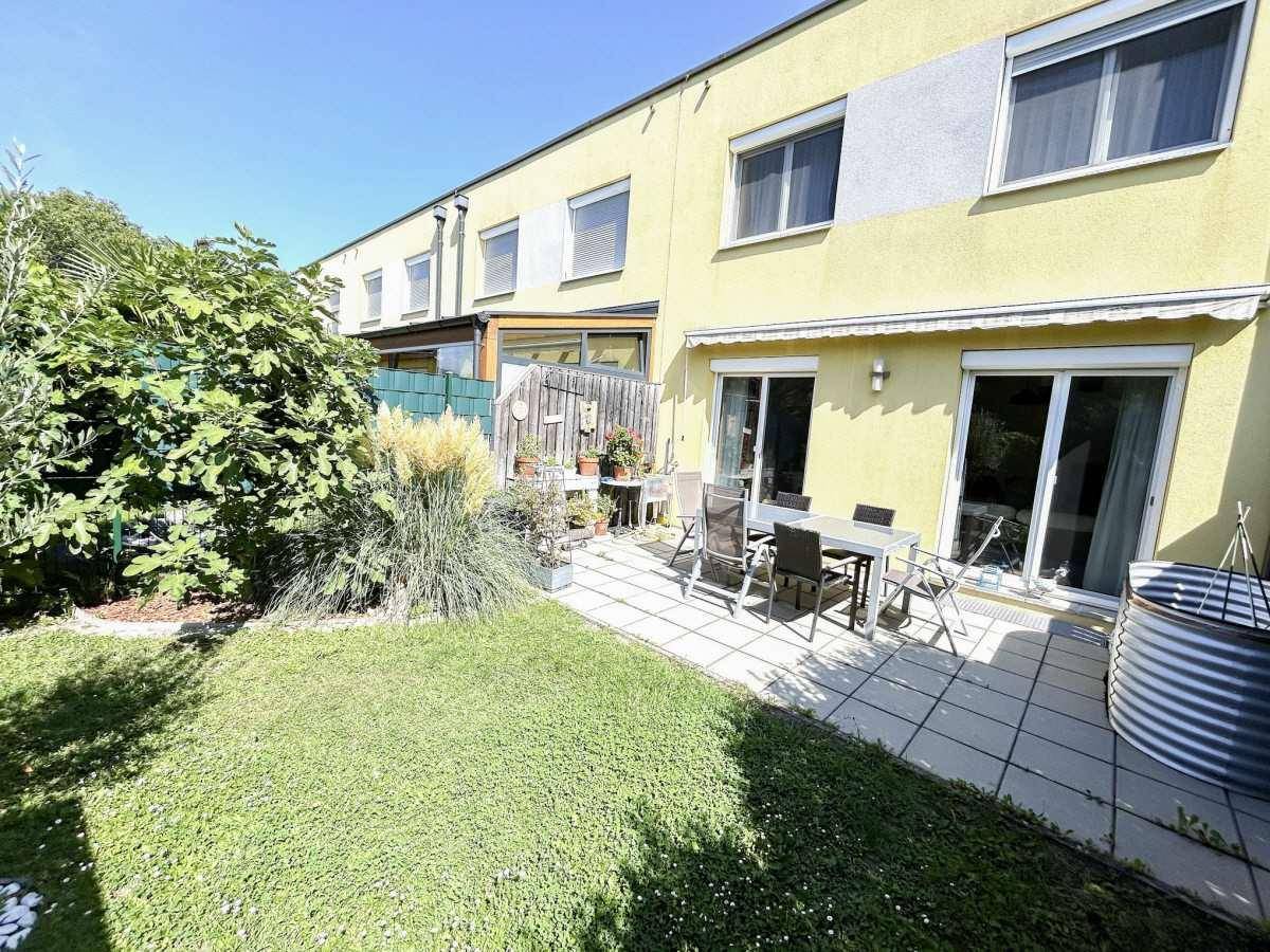 Sonniger Garten mit gepflasterter Terrasse, Rasenfläche und Zugang zum Haus, umgeben von Grün.