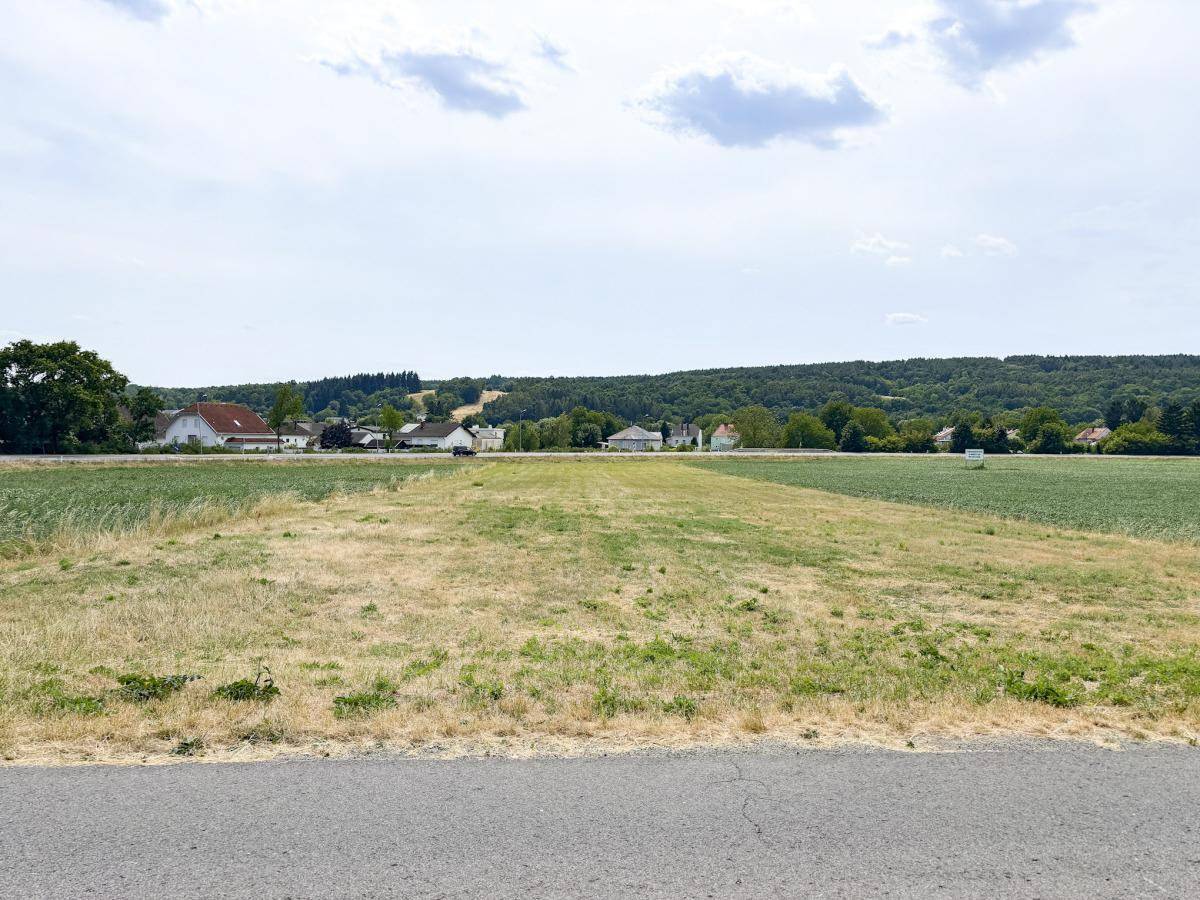 Blick über ein trockenes Feld auf eine ländliche Umgebung mit Häusern und bewaldeten Hügeln am Horizont.