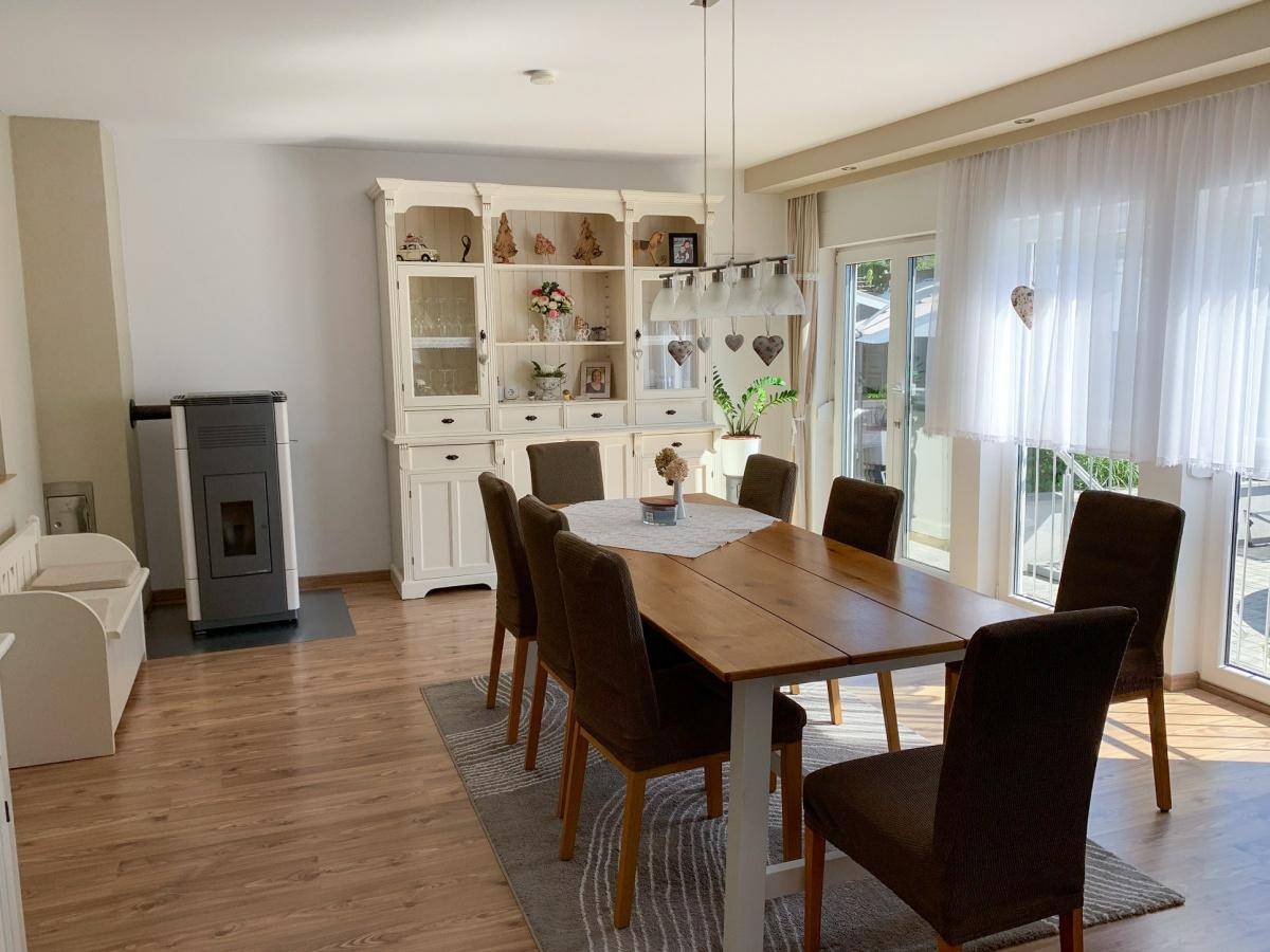 Heller Essbereich mit Holztisch, passenden Stühlen, Laminatboden und einem traditionellen Kachelofen.