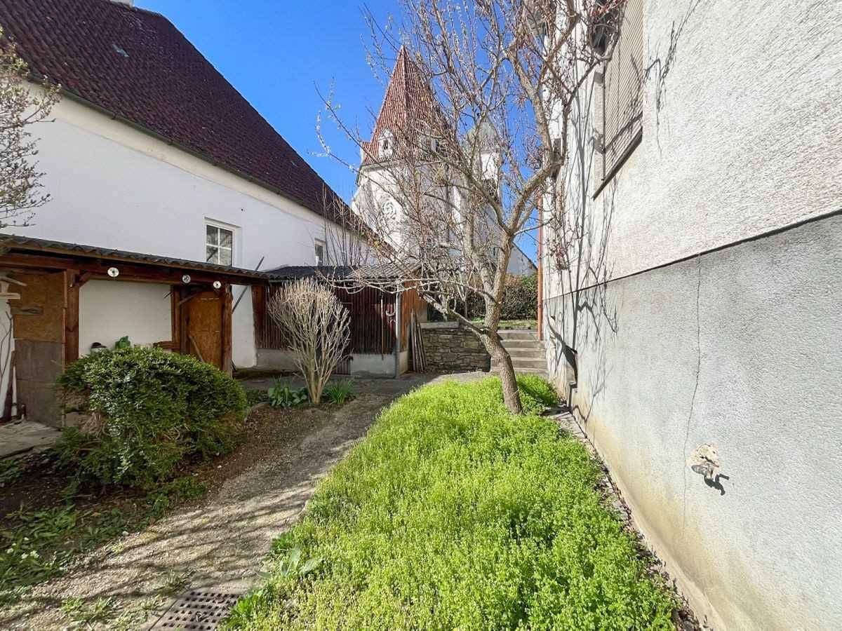 Schmaler Weg neben dem Haus mit grüner Bepflanzung und Blick auf einen Turm.