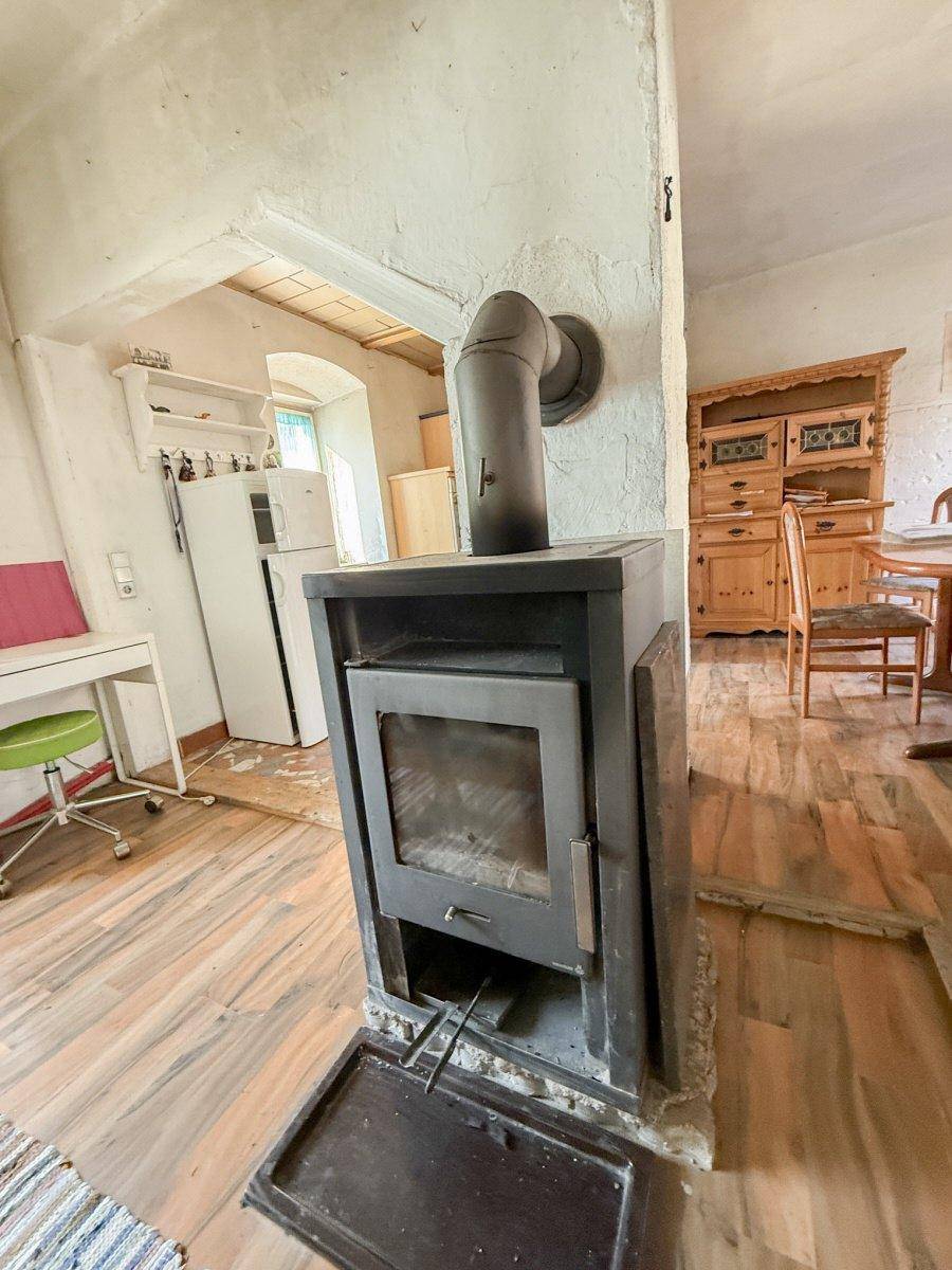 Gemütlicher Raum mit einem Holzofen, weißen Wänden und Holzboden, angrenzend an eine Küche.