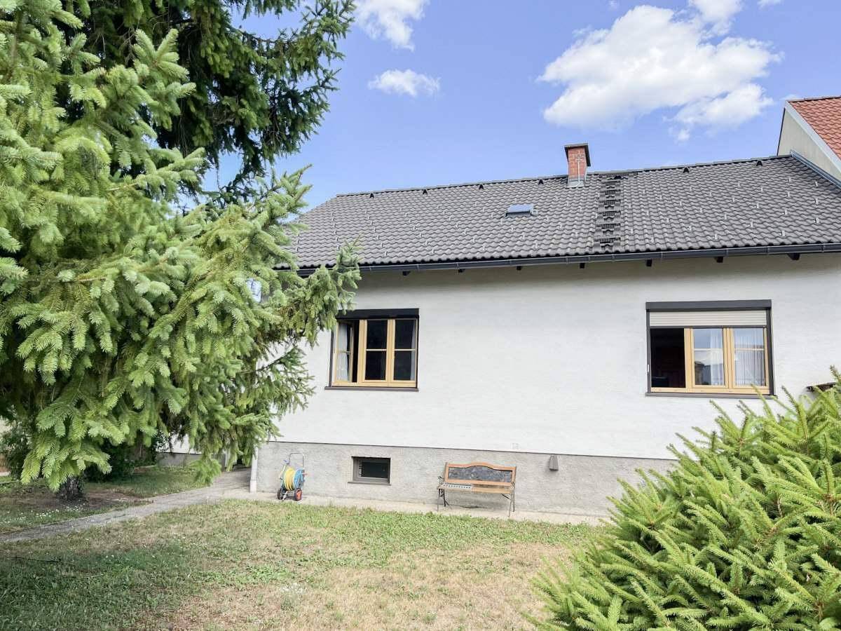 Seitenansicht des Hauses mit Garten, großen Nadelbäumen und einem kleinen Fenster im Kellerbereich.