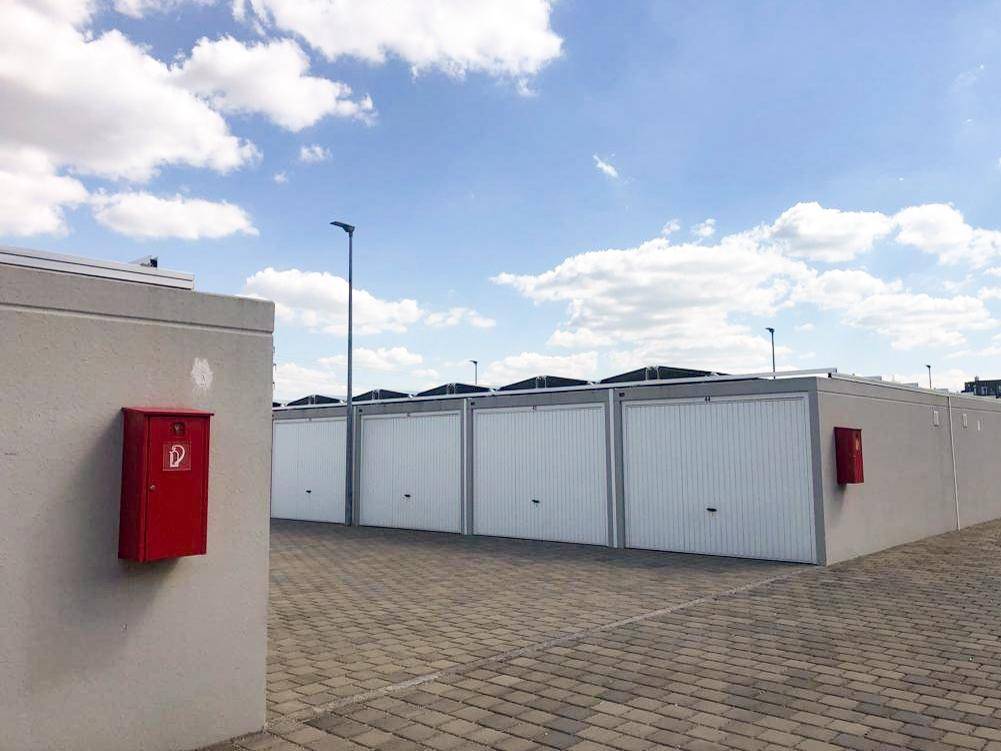 Reihe von weißen Garagen mit einem roten Feuerlöscherkasten an einer grauen Wand im Vordergrund.