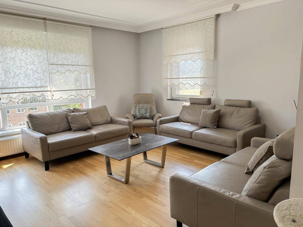 Helles Wohnzimmer mit beigen Ledersofas, Holzboden und großen Fenstern.