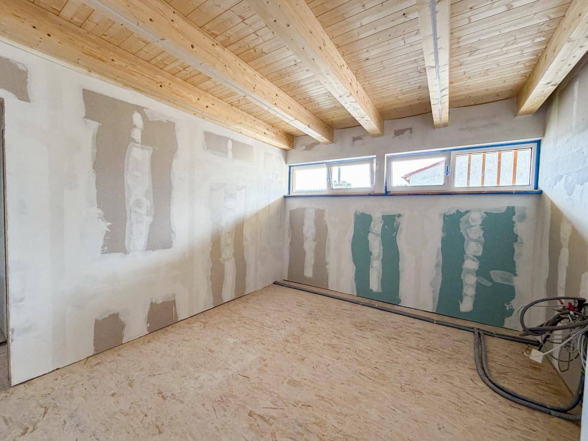Rohbau eines Zimmers mit Holzbalkendecke und horizontalen Fenstern, bereit für den Innenausbau.