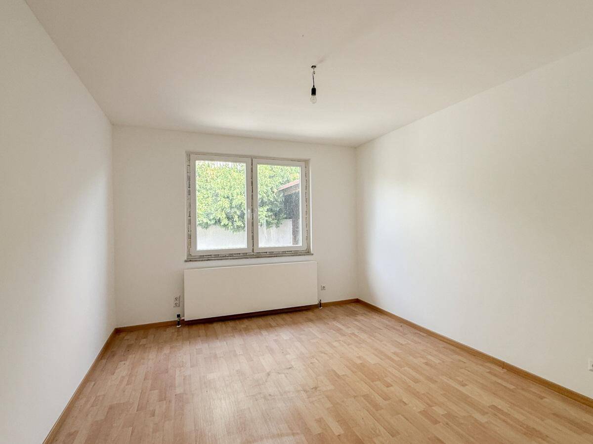 Geräumiges, leeres Zimmer mit Laminatboden und Fenster zum grünen Außenbereich.