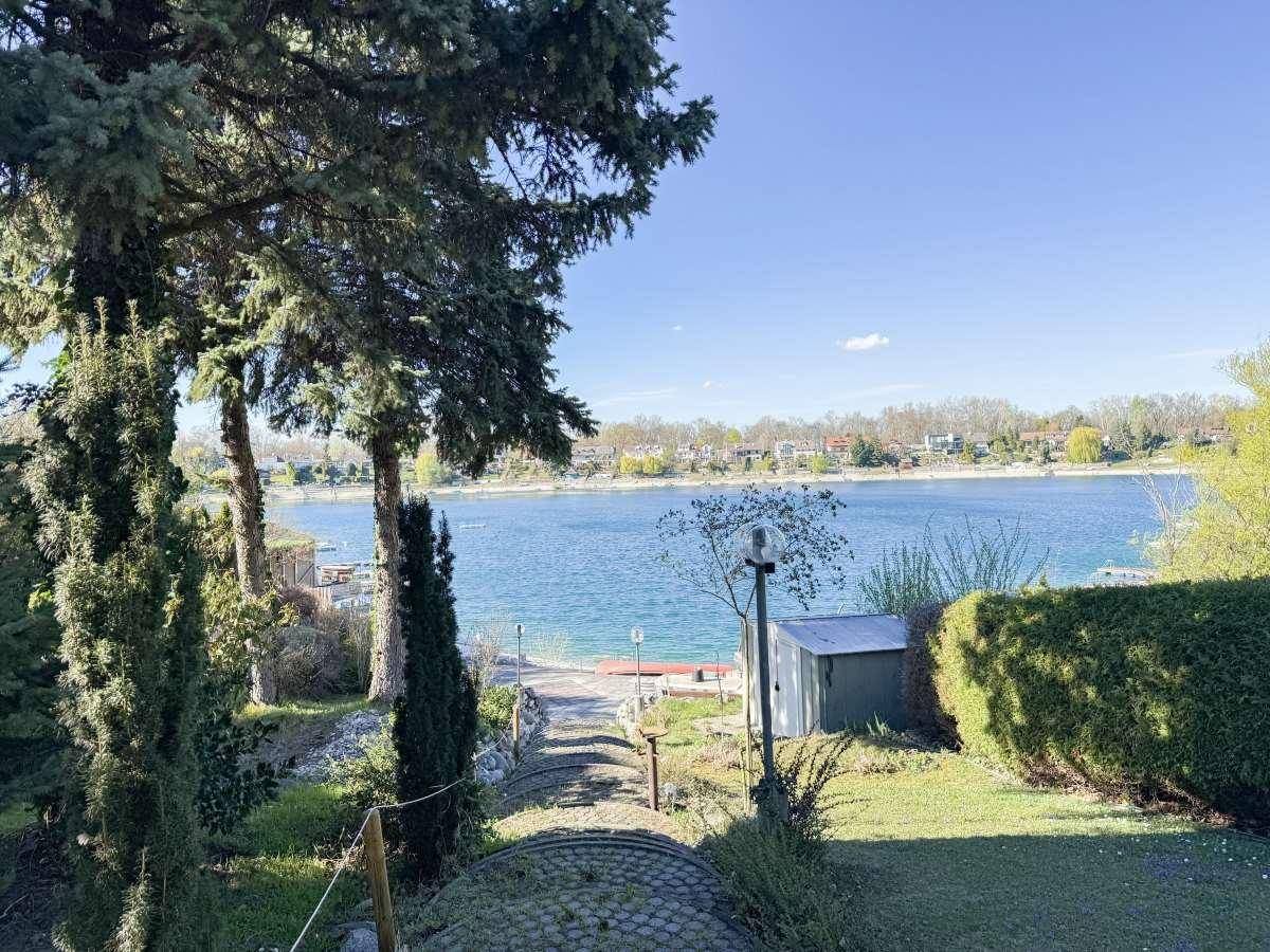 Blick vom Garten auf einen ruhigen See, umgeben von üppigem Grün und Bäumen unter blauem Himmel.
