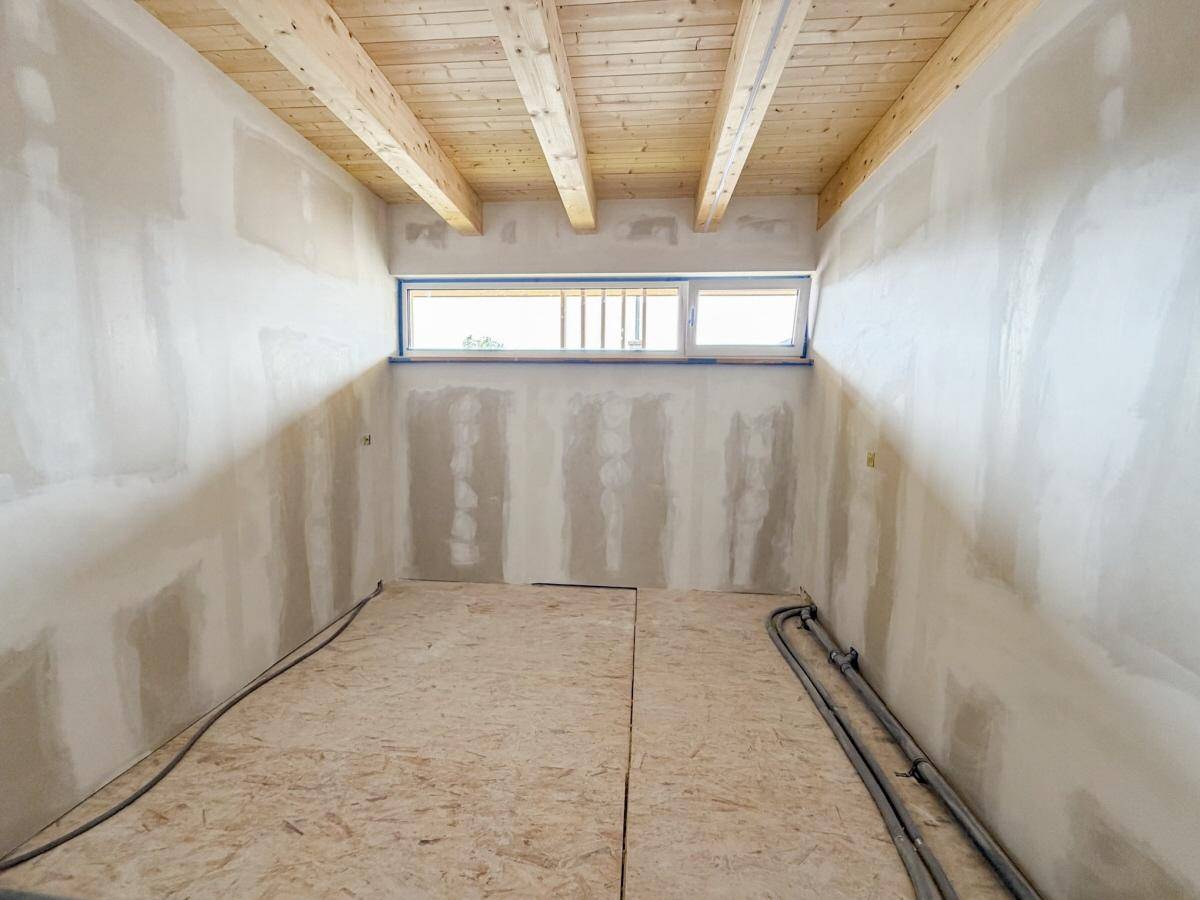 Innenraum im Rohbau mit Holzbalkendecke und einem breiten Fensterband, ideal für natürliches Licht.