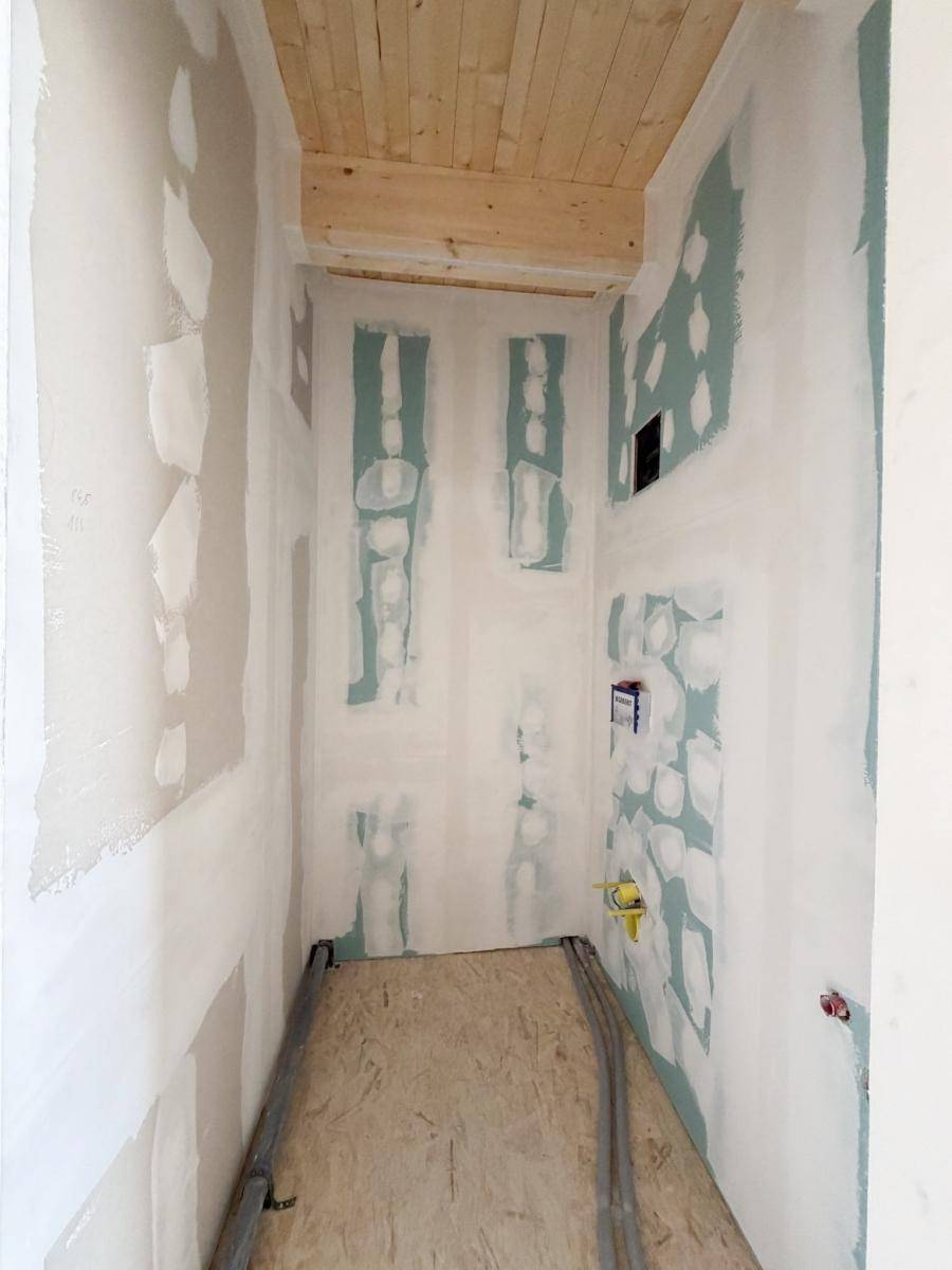 Schmaler Innenraum im Rohbau mit Holzbalkendecke und vorbereiteten Wänden, möglicherweise ein Flur.