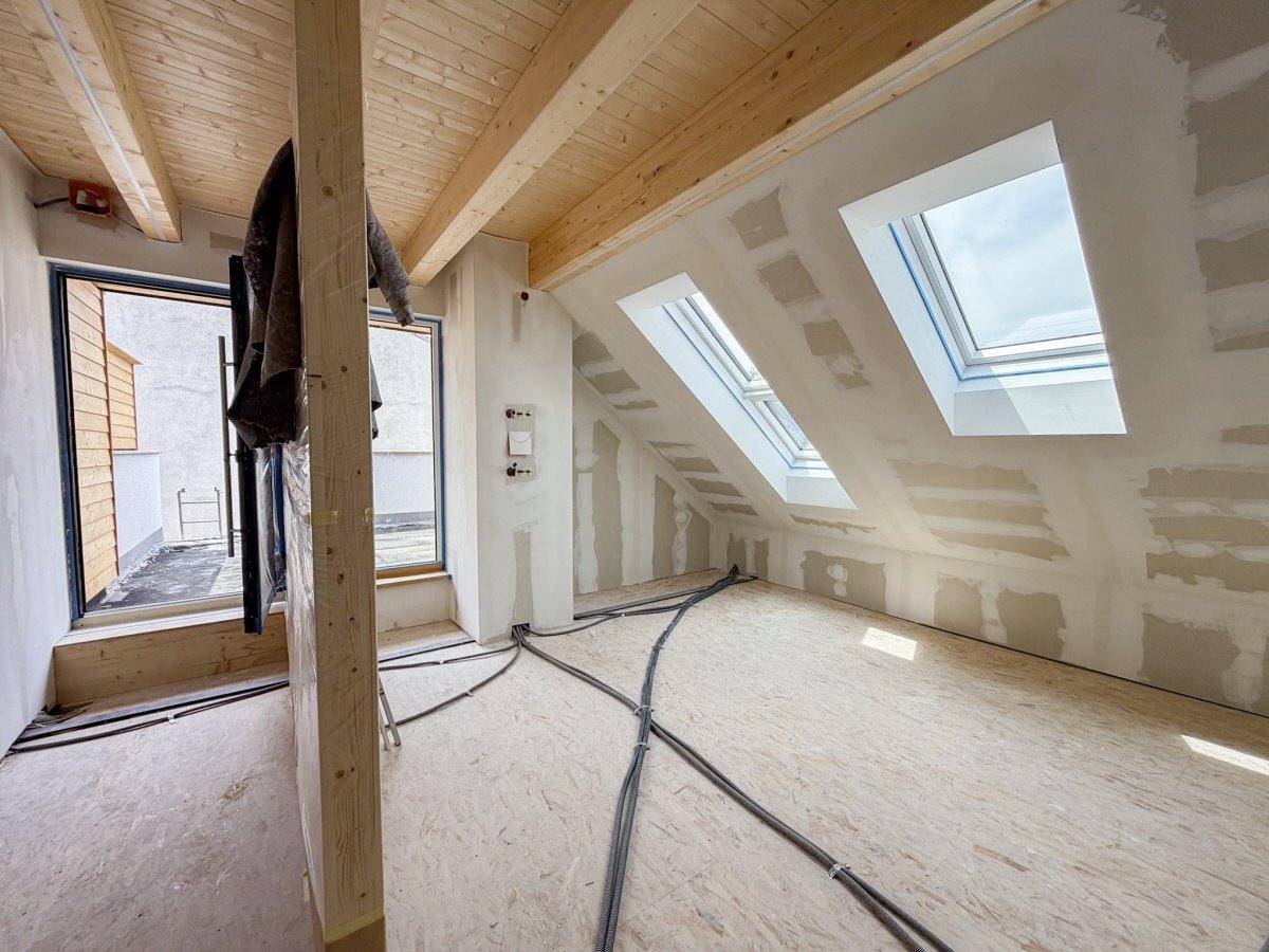 Rohbau eines Dachgeschossraumes mit Holzbalkendecke, mehreren Dachfenstern und Zugang zu einer Terrasse.