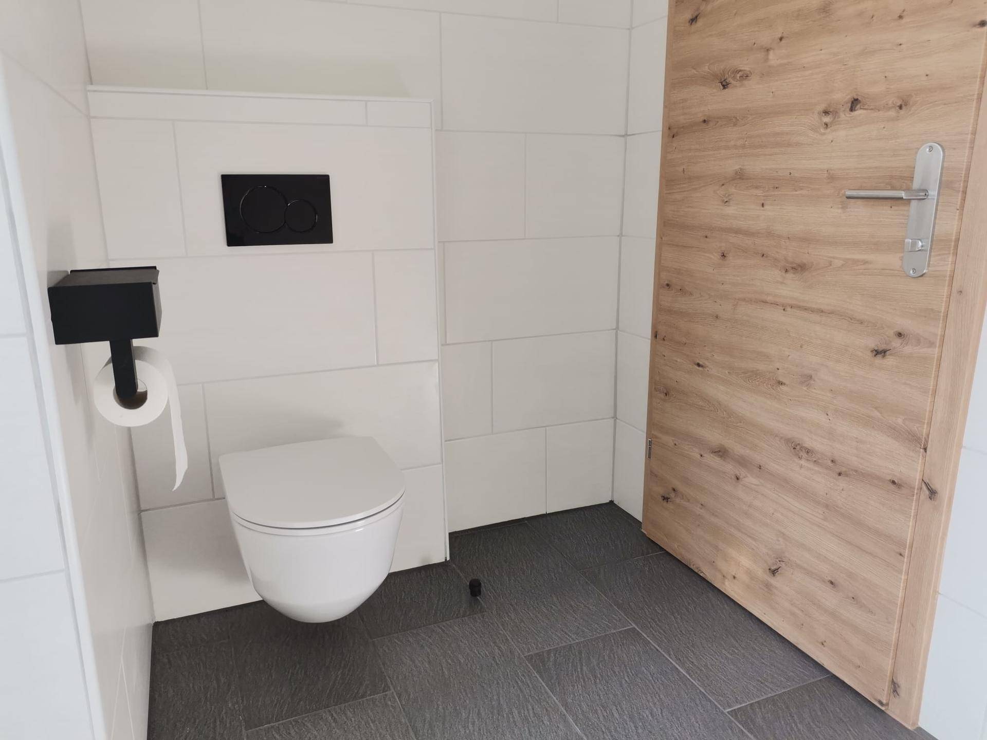 Separates WC mit wandhängender Toilette, weißen Wandfliesen und dunklem Fliesenboden.