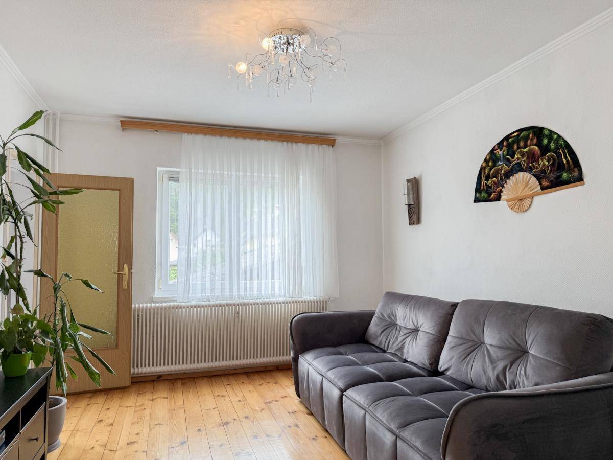 Wohnzimmer mit bequemer grauer Couch, Holzdielenboden und Fenster mit Blick ins Grüne.