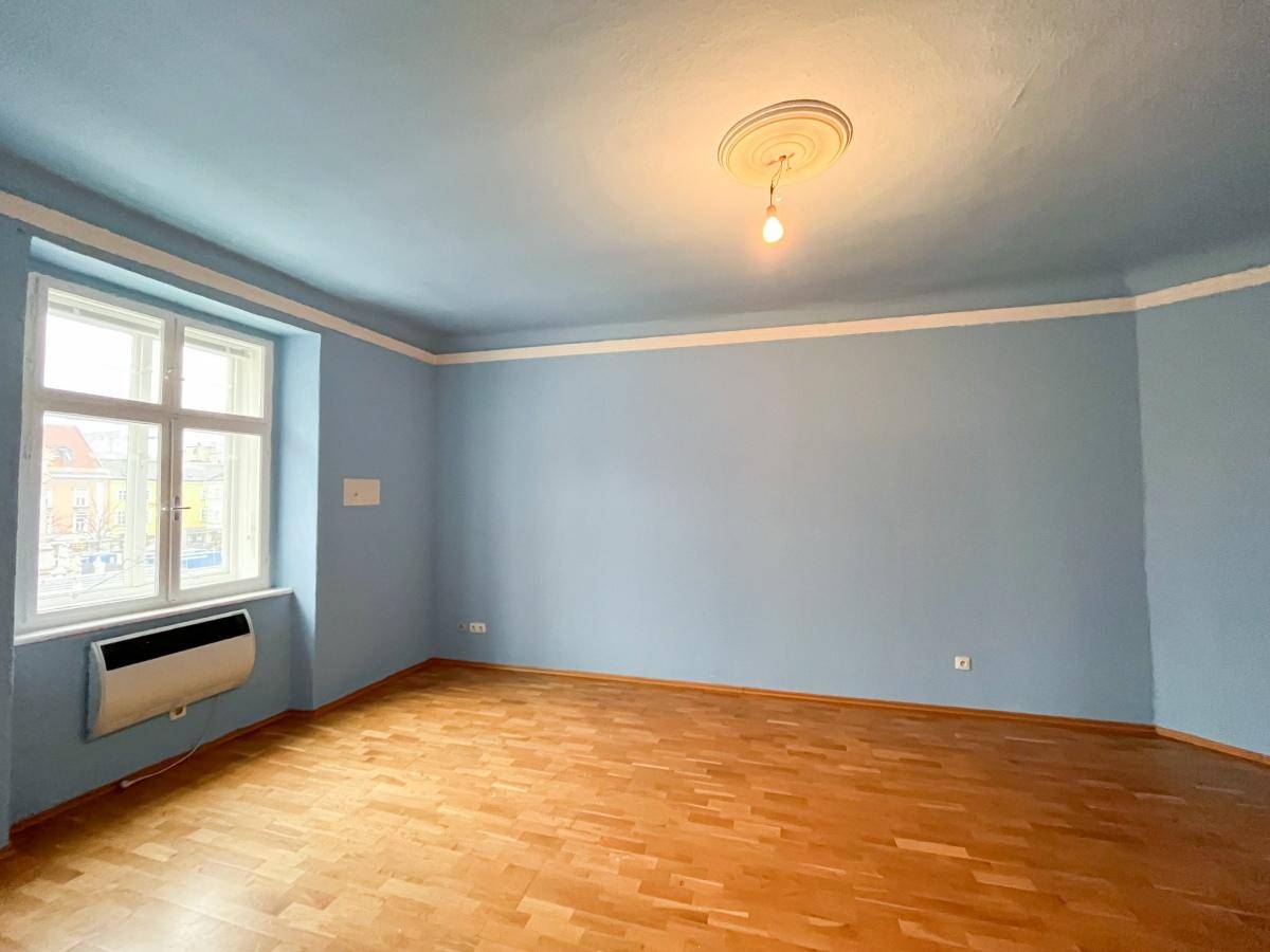 Geräumiges Zimmer mit hellblauen Wänden, Holzboden und einem Fenster, das viel Tageslicht hereinlässt.