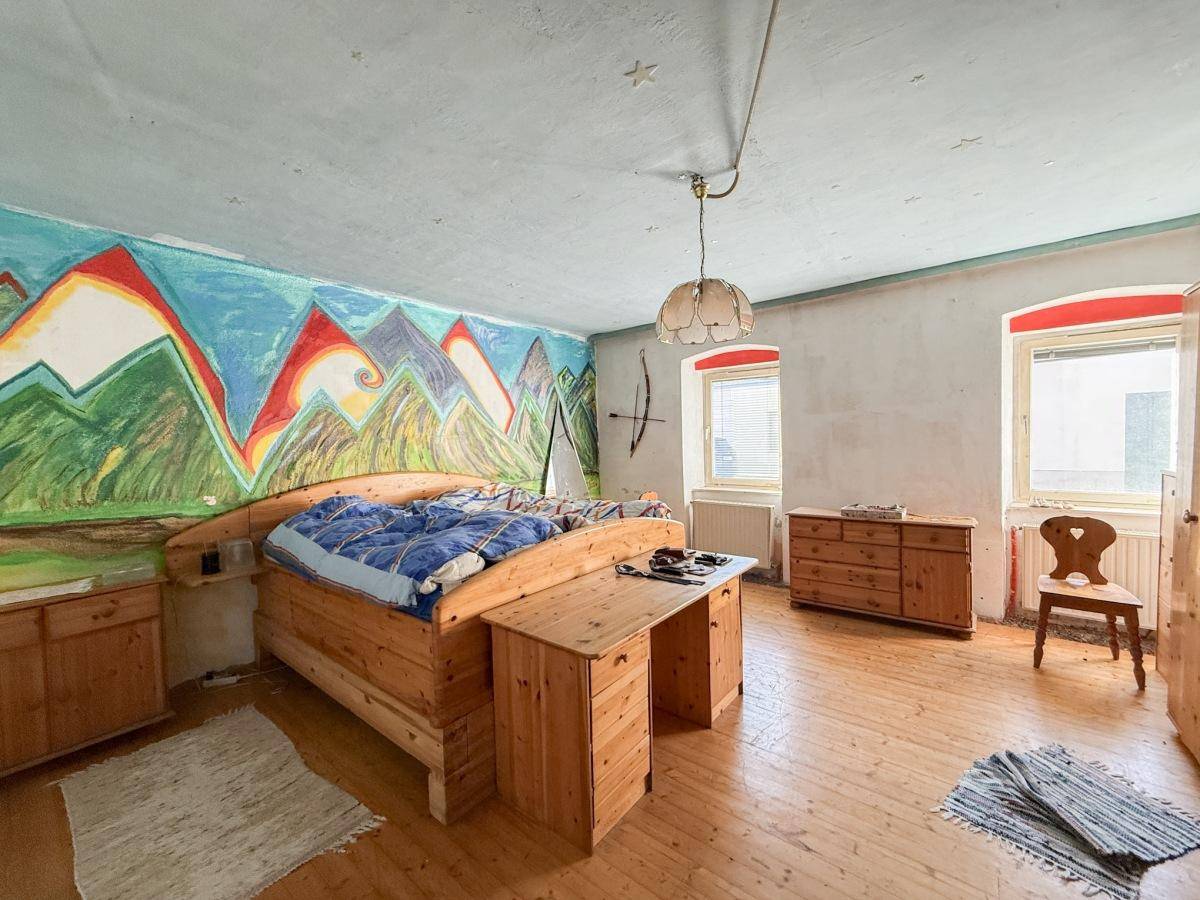 Schlafzimmer mit großem Holzbett, Holzkommode, Holzboden und einer farbenfrohen Wandmalerei.