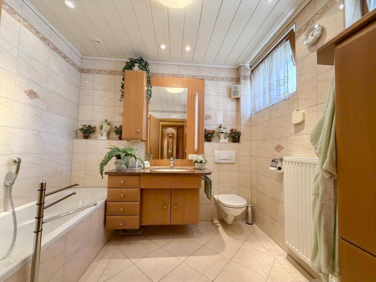 Geräumiges Badezimmer mit Badewanne, Waschtisch und hellen Fliesen, ausgestattet mit Holzmöbeln.