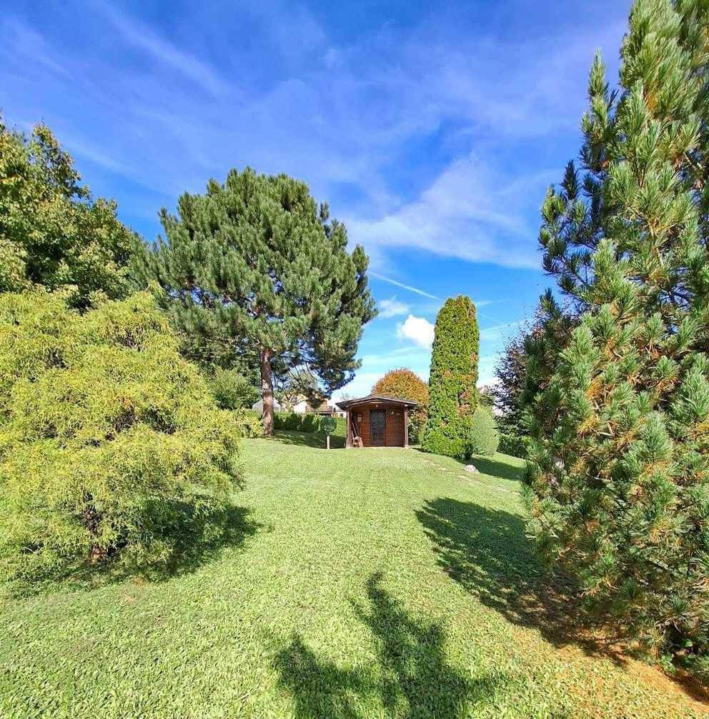 Großer, gepflegter Garten mit weitläufiger Rasenfläche, hohen Bäumen und einem Gartenhaus unter blauem Himmel.