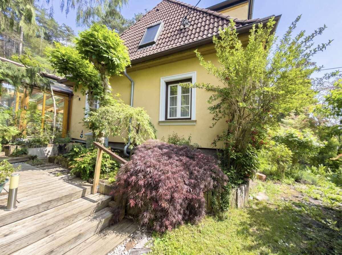 Seitenansicht des Hauses mit Holztreppe und üppiger Bepflanzung im Vorgartenbereich.