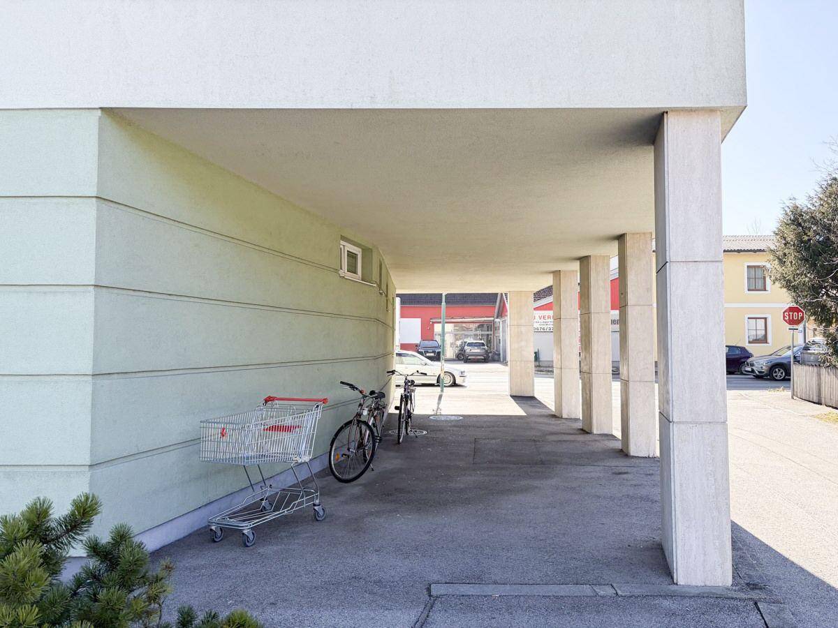 Überdachter Bereich unter dem Gebäude mit Säulen, Fahrrädern und einem Einkaufswagen.