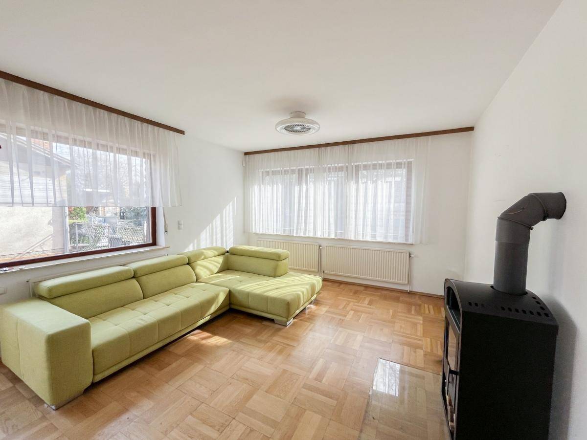 Gemütliches Wohnzimmer mit hellem Parkettboden, grünem Ecksofa und Holzofen.