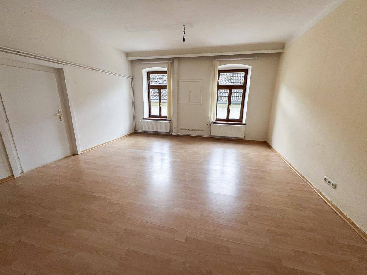 Leeres Wohnzimmer mit Laminatboden, zwei Fenstern mit Holzrahmen und weiß gestrichenen Wänden.