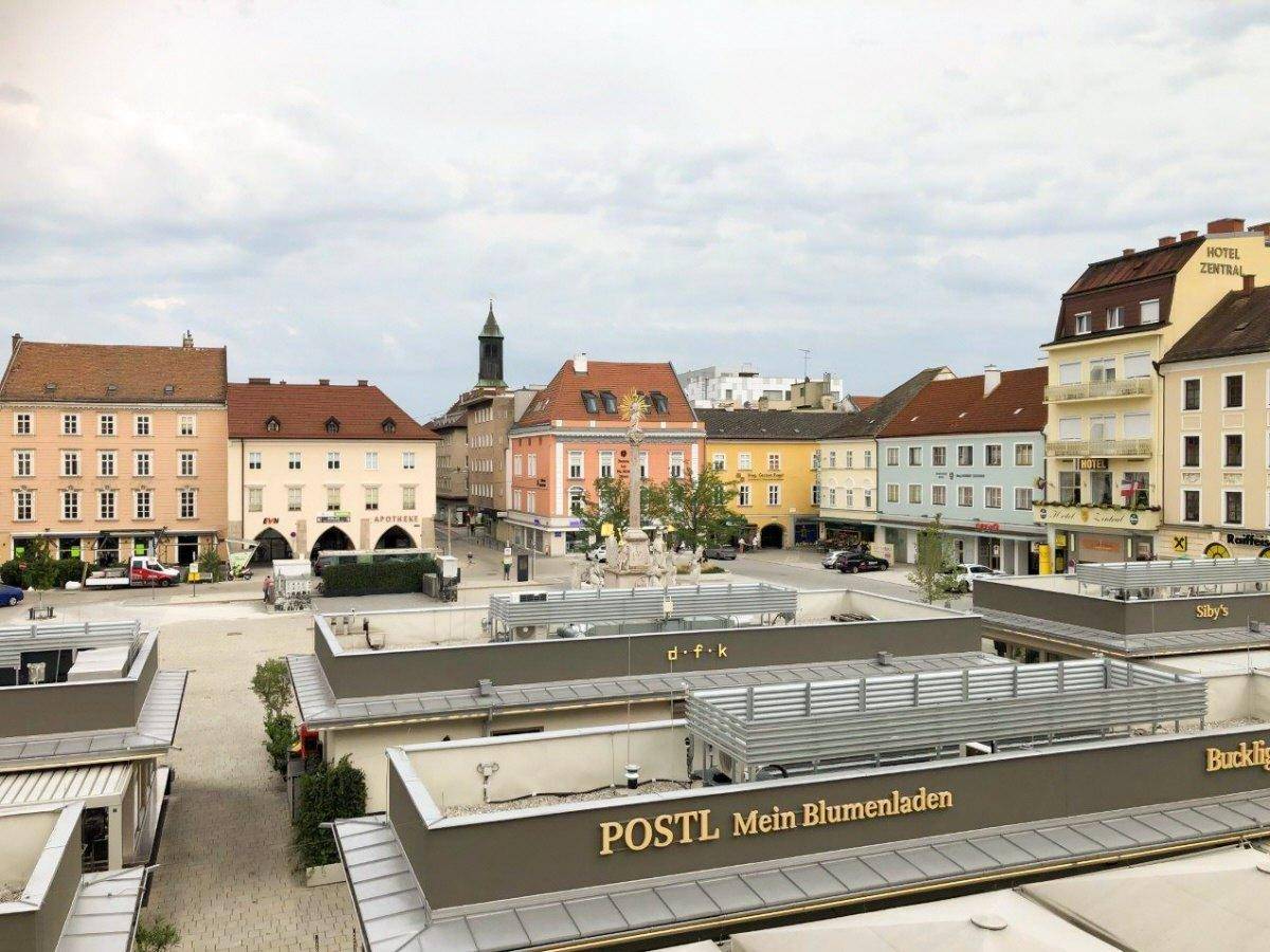 Weitläufiger Blick über einen Stadtplatz mit verschiedenen Gebäuden und Geschäften unter einem bewölkten Himmel.