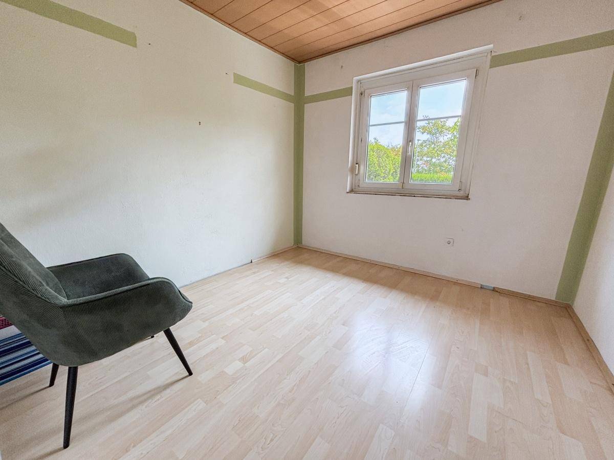 Schlafzimmer mit hellem Laminatboden, weiß gestrichenen Wänden und Fensterblick ins Grüne.