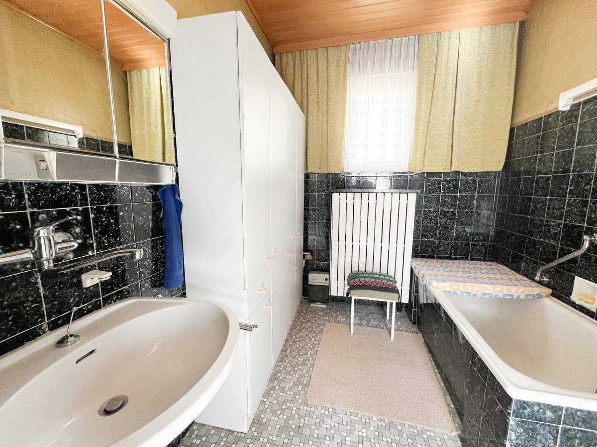 Badezimmer mit Badewanne, großem Waschbecken, Spiegelschrank und dunklen Fliesen.