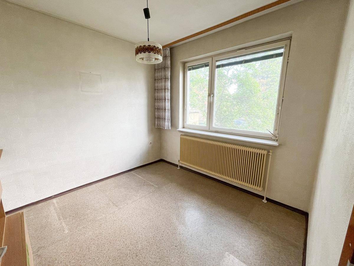 Leeres Zimmer mit großem Fenster, Heizkörper und einfacher Deckenleuchte.