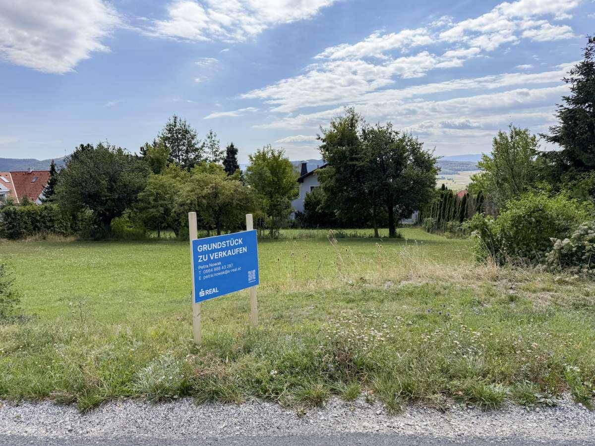 Grundstück mit "Zu verkaufen"-Schild, umgeben von grüner Landschaft und vereinzelten Häusern.