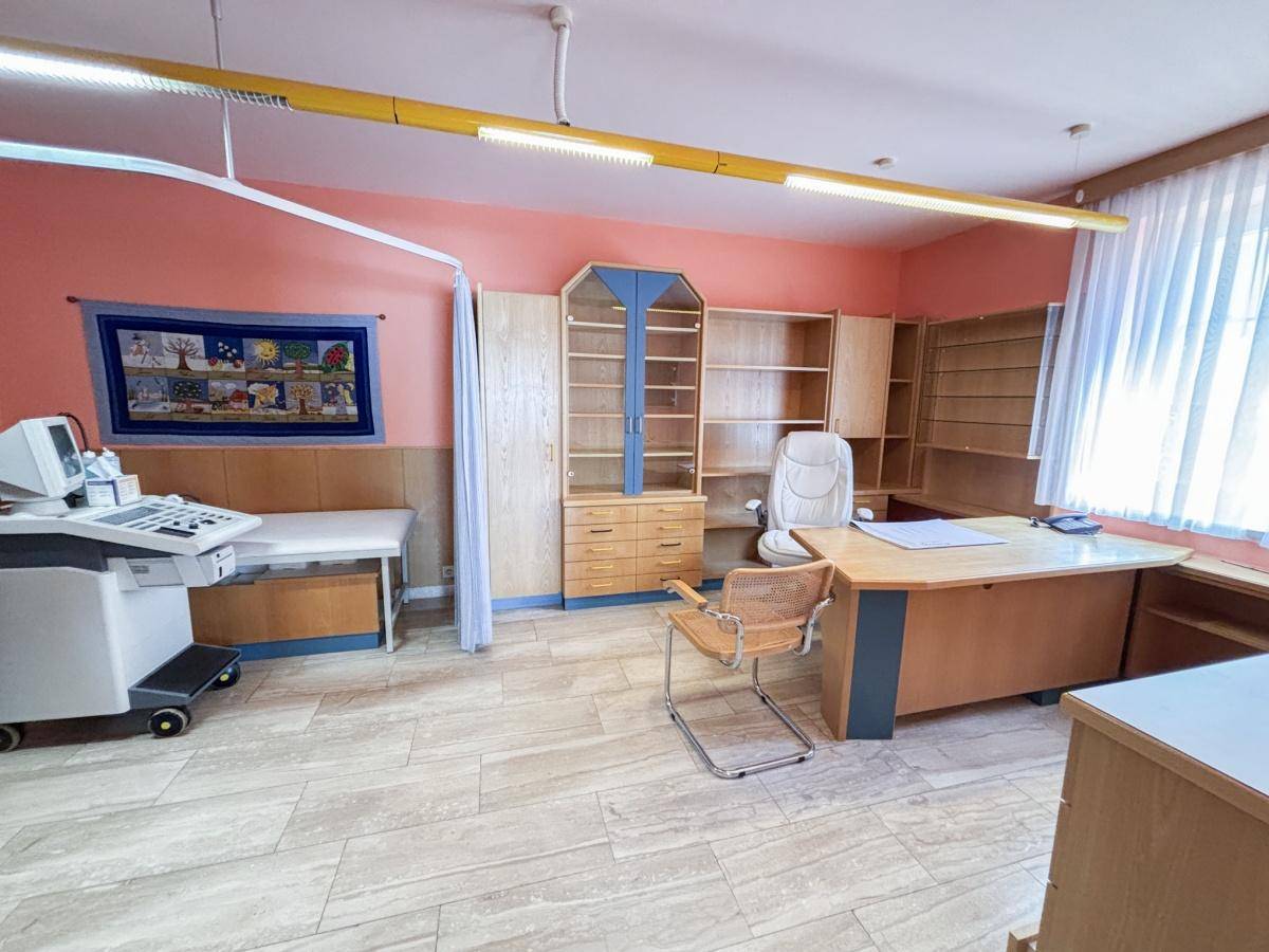 Behandlungszimmer mit medizinischen Geräten, Schreibtisch und Schränken, in einem hellen Rosaton gestrichen.