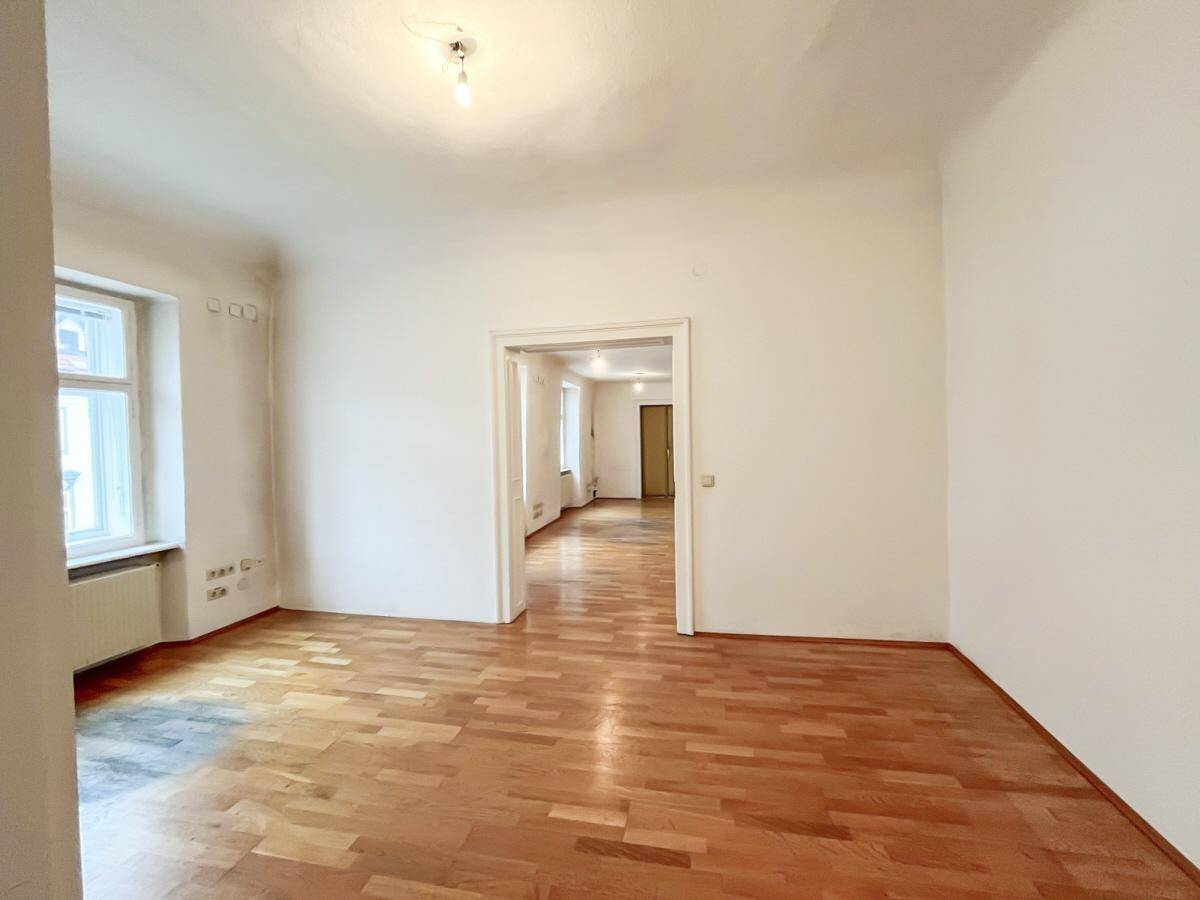 Helles Zimmer mit Holzboden und weißen Wänden, das durch eine offene Tür mit einem weiteren Raum verbunden ist.