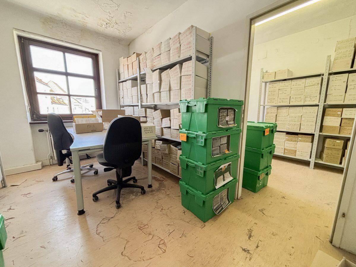 Büro- und Archivraum mit einem Schreibtisch, Bürostühlen und Regalen voller Kartons.