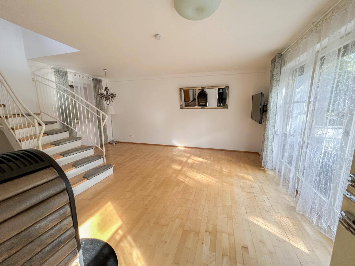 Geräumiges Wohnzimmer mit hellem Holzboden, Treppe und großen Fenstern.