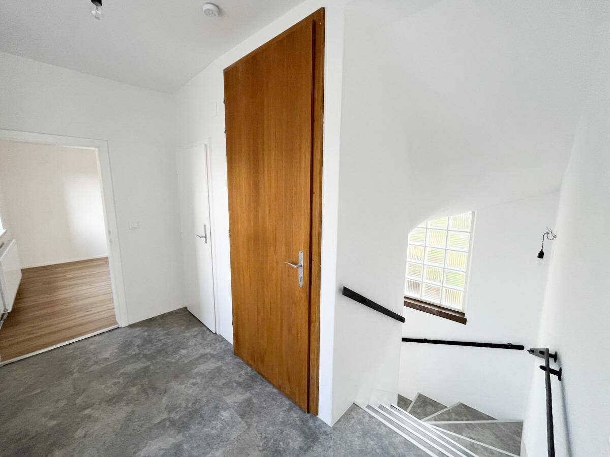 Flur mit Treppenaufgang, Holztür und einem Fenster aus Glasbausteinen.