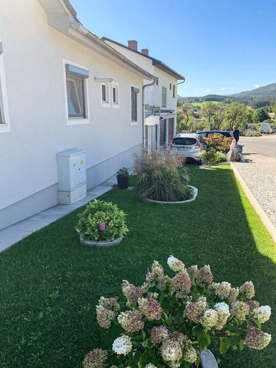 Gepflegter Vorgarten mit Rasenfläche, Blumenbeeten und Blick auf die umliegende Landschaft.