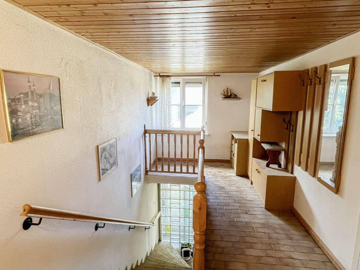 Oberer Flurbereich mit Holzgeländer, Fliesenboden und Einbauschränken, hell durch Fenster.