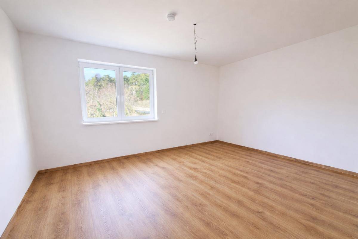 Leeres Zimmer mit hellem Laminatboden und einem Fenster, das viel Tageslicht hereinlässt.