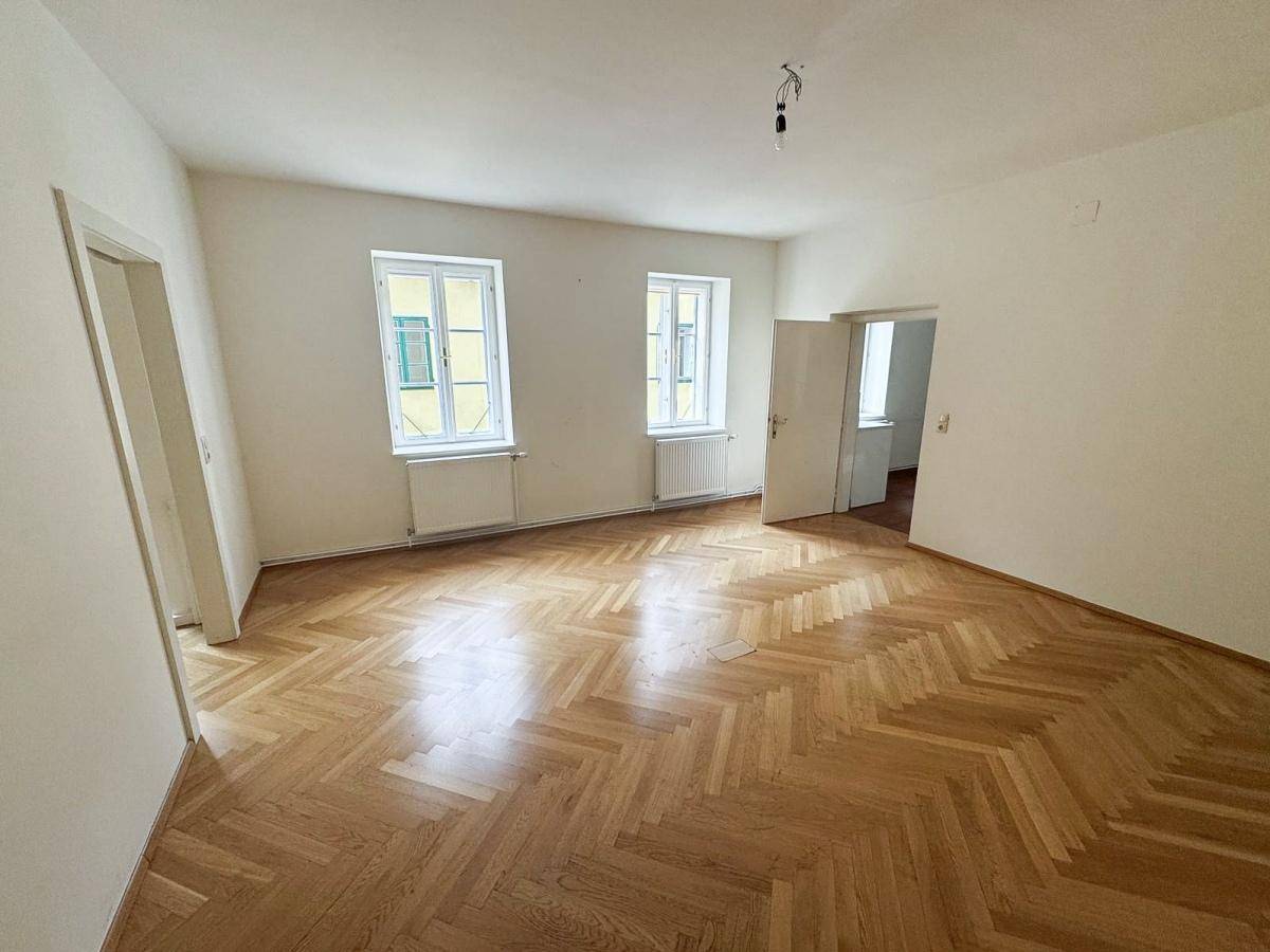 Helles Wohnzimmer mit Fischgrätparkett, zwei großen Fenstern und Zugang zu weiteren Räumen.